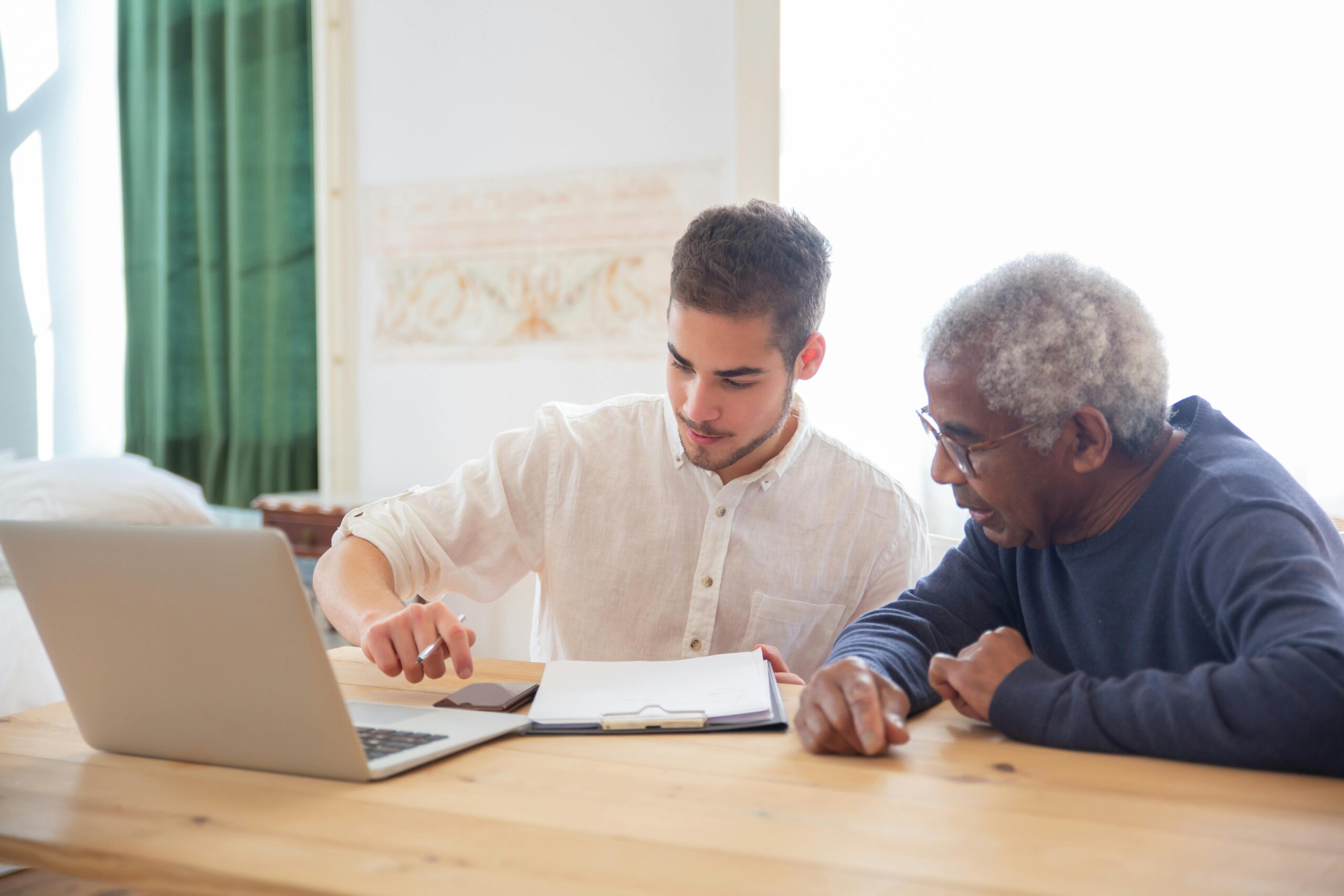 Young man teaches elderly gentleman to use technology at home.